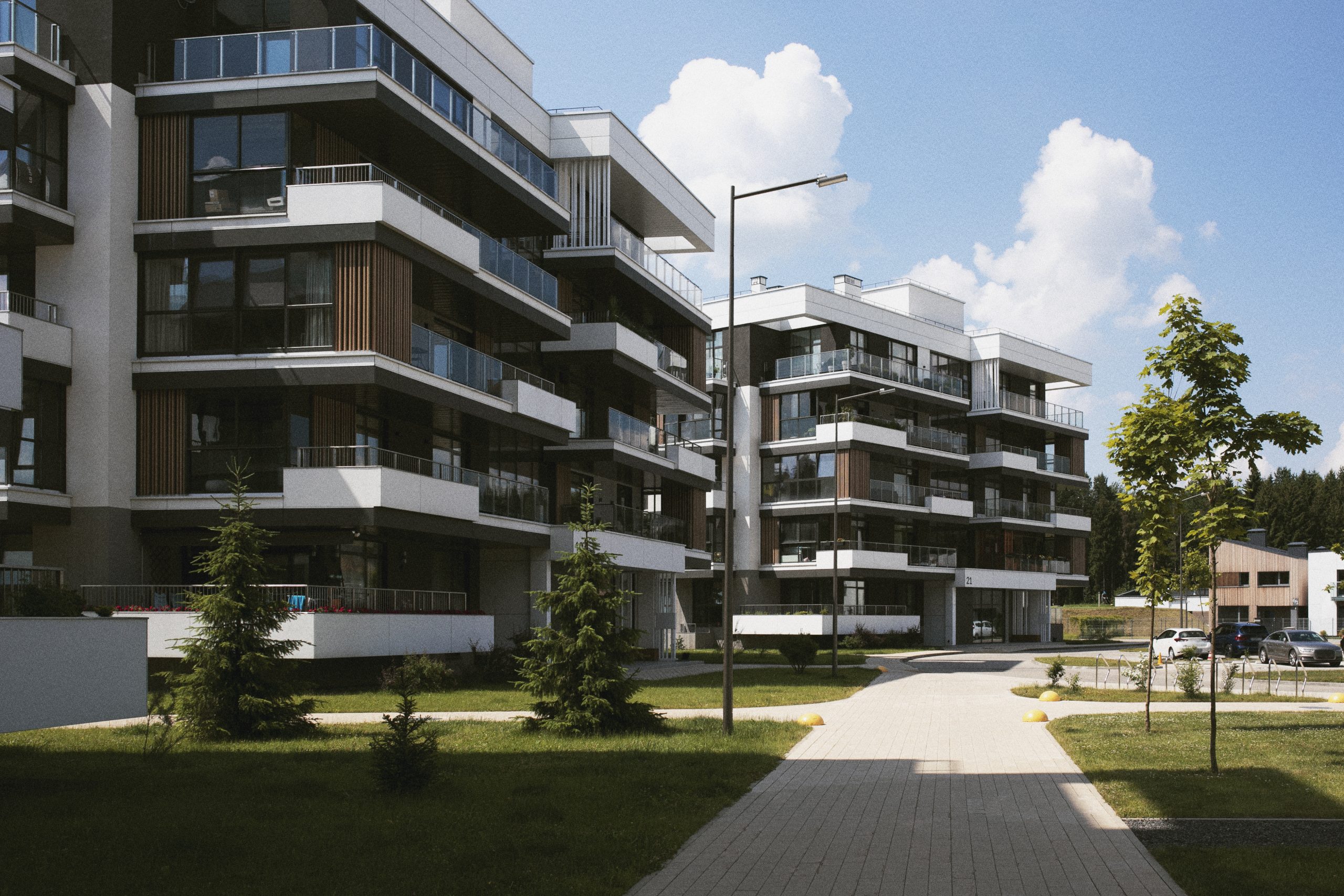 Dwa nowoczesne, kilkupiętrowe budynki apartamentowe z białymi i drewnianymi elewacjami oraz licznymi balkonami, otoczone zadbanym trawnikiem i ścieżką.