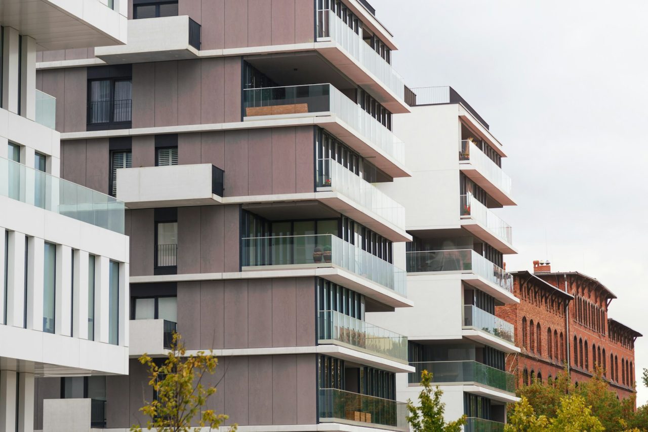 Rząd nowoczesnych budynków apartamentowych z balkonami ze szklanymi barierkami, sąsiadujący ze starszą kamienicą z czerwonej cegły.